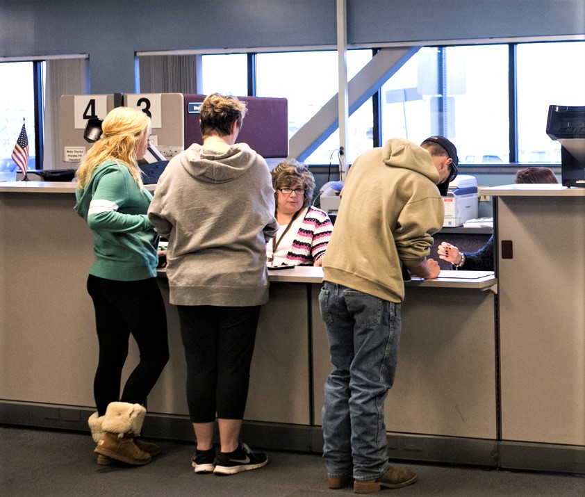Three customers stand at a Driver and Vehicle Services counter while two employees assist them.